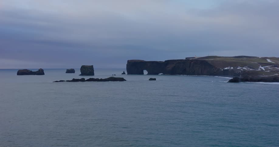 A dramatic wide shot of the iconic Dyrhólaey rock arch and sea stacks along the Icelandic coast on a moody, overcast day