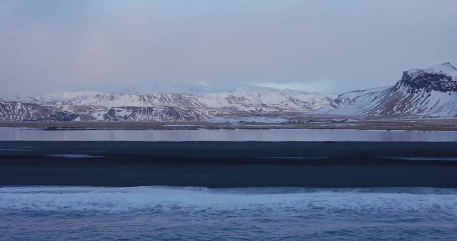 A serene, wide shot of gentle ocean waves lapping against a black sand beach in Iceland, with a vast, snow-covered mountain range in the background