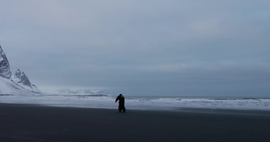 A lone figure in a dark cloak walks along a desolate black sand beach next to snow-covered mountains and a cold ocean