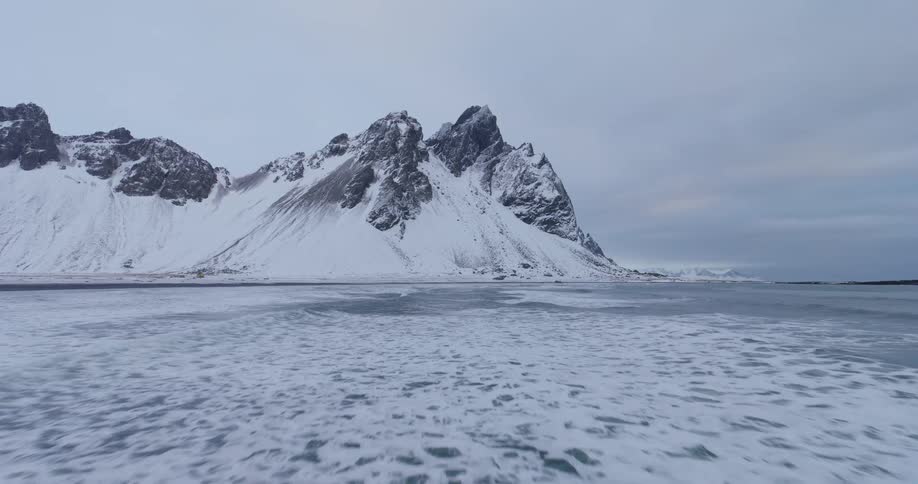 An epic slow-motion aerial shot glides over rolling waves towards the dramatic snow-covered Vestrahorn mountain range in Iceland