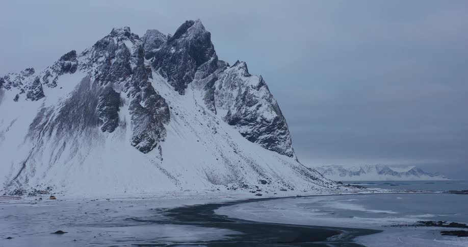 A majestic, snow-covered Vestrahorn mountain towers over a frozen black sand beach and the cold North Atlantic ocean in Iceland