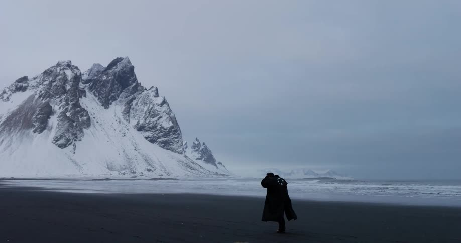 A solitary figure in a long dark coat walks along a vast black sand beach, battling the wind with epic snow-covered mountains and the ocean in the background