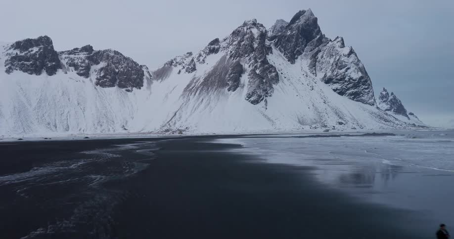 A lone figure walks along a vast black sand beach at the base of the epic snow-covered Vestrahorn mountain in Iceland