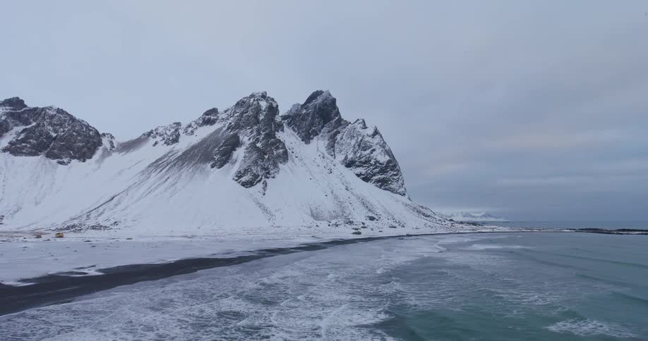 A majestic snow-covered mountain looms over a black sand beach as icy waves crash ashore on a cold, overcast day