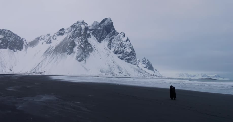 A person in a long black coat walks along a vast black sand beach with dramatic snow-covered mountains in the background