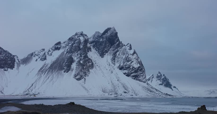 Aerial view of the majestic, snow-covered Vestrahorn mountain range towering over a frozen black sand beach in Iceland
