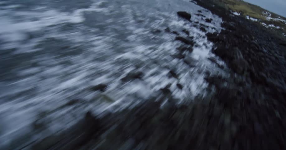A fast-paced, low-angle FPV drone flight speeds over the crashing waves along a rugged and cold rocky shoreline
