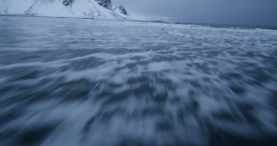 A fast-paced, low-angle drone shot skims over the surface of the ocean as waves recede from a snowy beach with mountains in the background