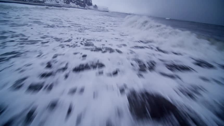 A dynamic drone shot soars low and fast over the turbulent, foamy waves of a cold ocean crashing against a snowy black sand shoreline
