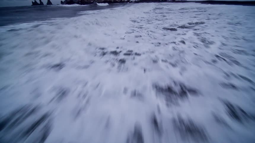 A dynamic point-of-view aerial shot skims just above the churning white surf as it washes onto a dramatic black sand beach in winter