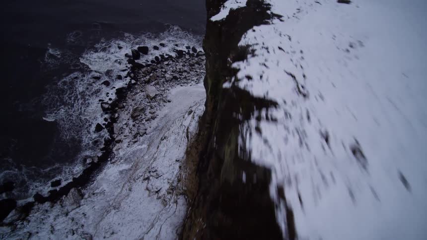 A moody aerial view reveals a vast, snow-dusted cliff face towering over a stormy ocean coastline on a winter day
