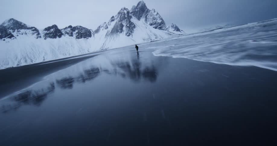 A dramatic low-angle tracking shot follows a lone runner along the shoreline of a black sand beach with epic snow-covered mountains in the background