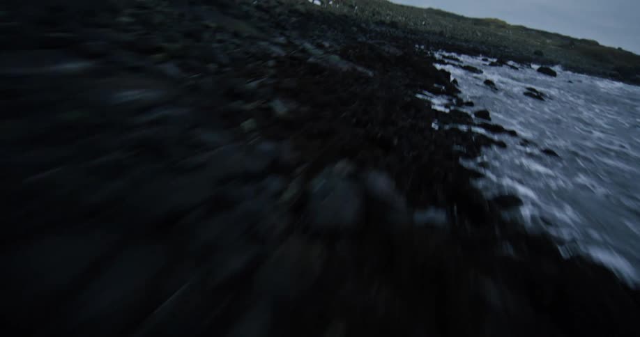 A fast-paced, low-level aerial shot speeds over a dramatic and rugged black rock shoreline as waves crash on a cold, overcast day