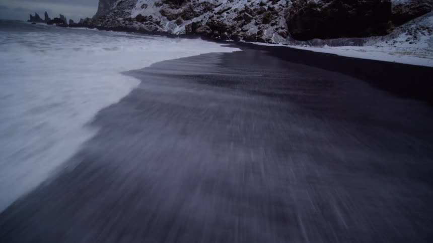 A stunning low-angle, slow-motion shot glides along the foamy surf as it recedes from a black sand beach next to snow-covered cliffs