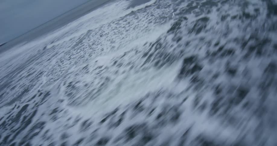 A high-speed first-person view races just above the turbulent and foamy ocean surface on a cloudy day
