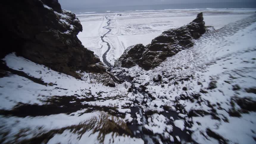 A dramatic first-person view flies over the edge of a snowy cliff, revealing a winding river flowing through a vast winter valley towards the ocean