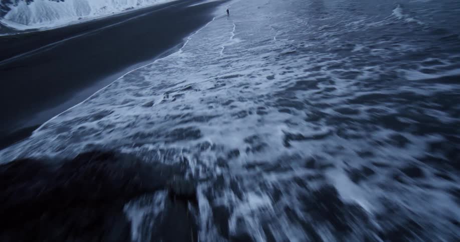 Exhilarating aerial footage follows a lone runner along a desolate black sand beach with massive snow-covered mountains in the background