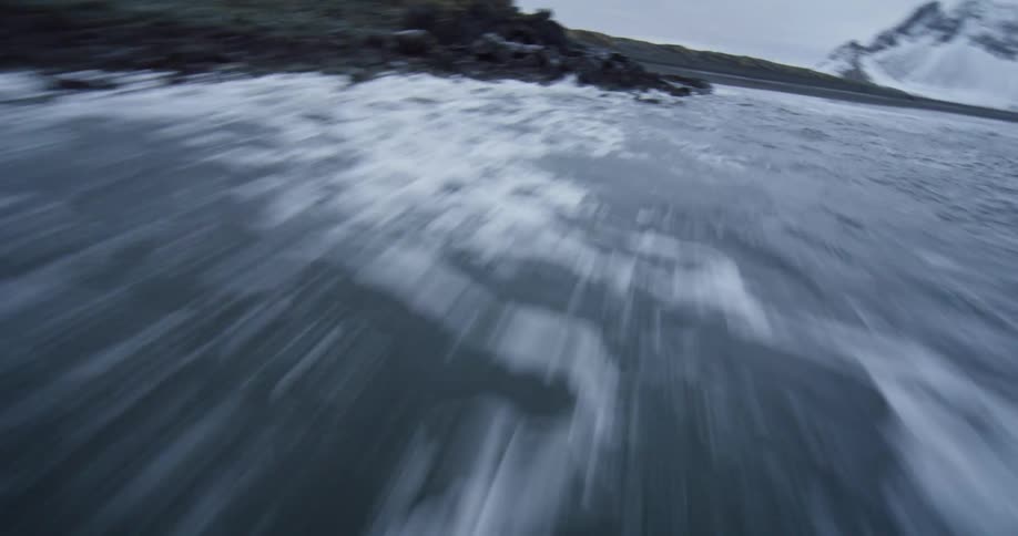 Dramatic FPV drone shot soars over the churning ocean waves crashing on a black volcanic rock shore with snowy mountains in the background