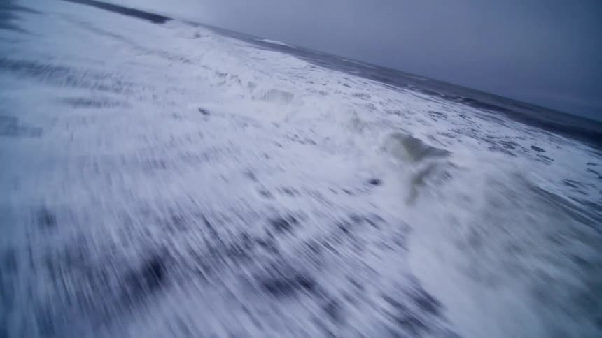 An immersive slow-motion view captures the powerful white foam of ocean waves washing over a dramatic black sand beach
