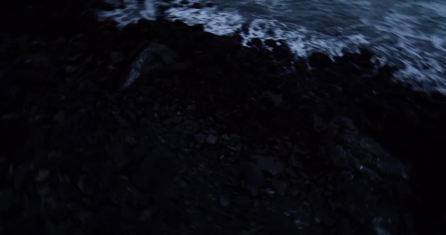 A thrilling, fast-paced aerial view skims over dark, choppy waters crashing onto a rocky, snow-dusted coast at twilight