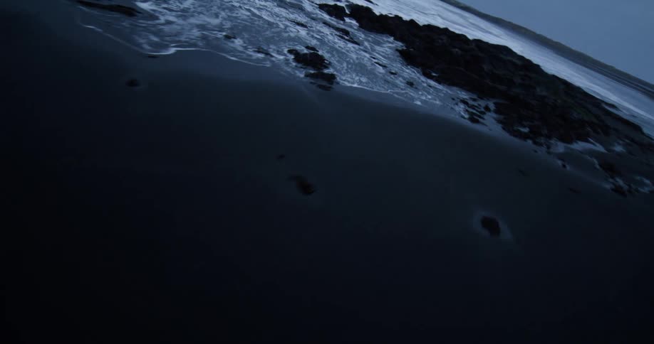 An intense, fast-moving shot skims low over a cold, black volcanic beach and rocky coastline as the ocean's white foam rushes in