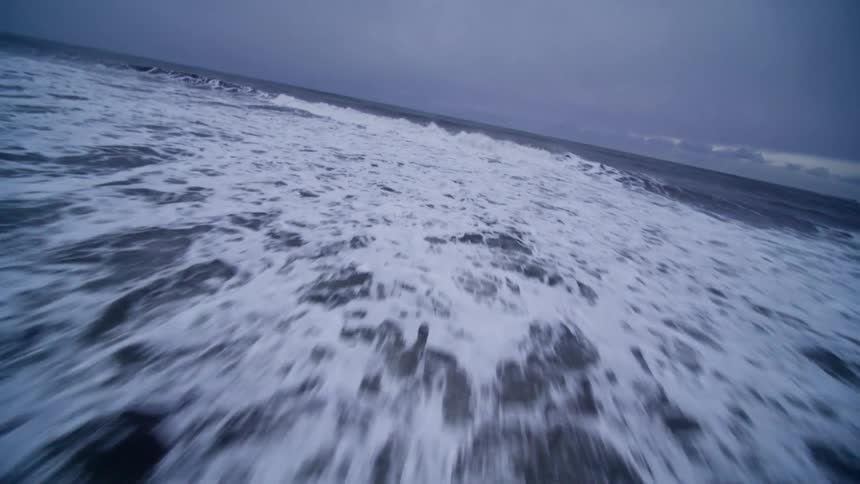 A dramatic low-angle shot glides from a snowy landscape across a black sand beach and into the powerful ocean waves under an overcast sky