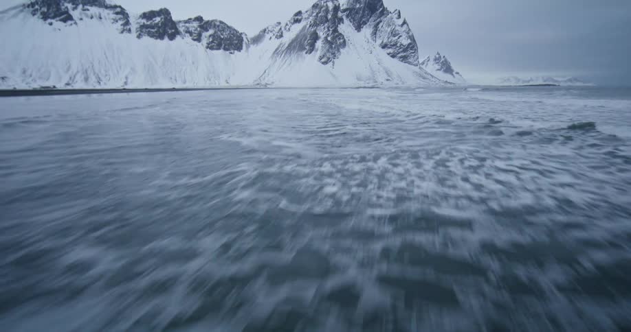 A low-angle shot follows the tide receding from a black sand beach with dramatic snow-covered mountains in the background