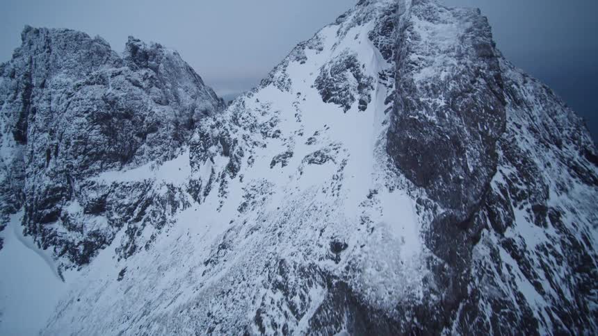 A breathtaking aerial shot ascends a rugged, snow-covered mountain peak to reveal the vast ocean on an overcast day