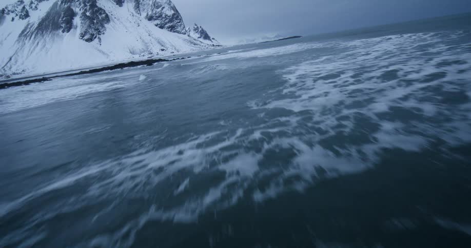 A thrilling low-angle drone shot skims across the cold ocean waves towards a dramatic, snow-covered mountain coastline