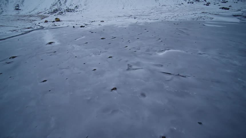 A slow aerial pullback reveals a desolate, snow-covered black sand beach at the base of a majestic mountain