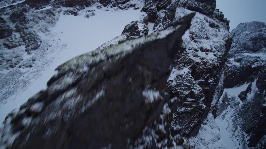 A dramatic aerial ascent along a rugged, snow-covered mountain cliff face in winter