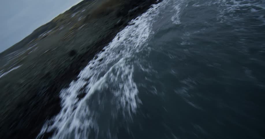 A dizzying, fast-paced aerial view flies over the chaotic ocean waves crashing against a snowy, black sand beach