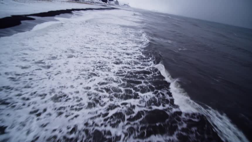 An exhilarating FPV drone shot flies down a snow-covered cliff and skims over the crashing waves on a black sand beach
