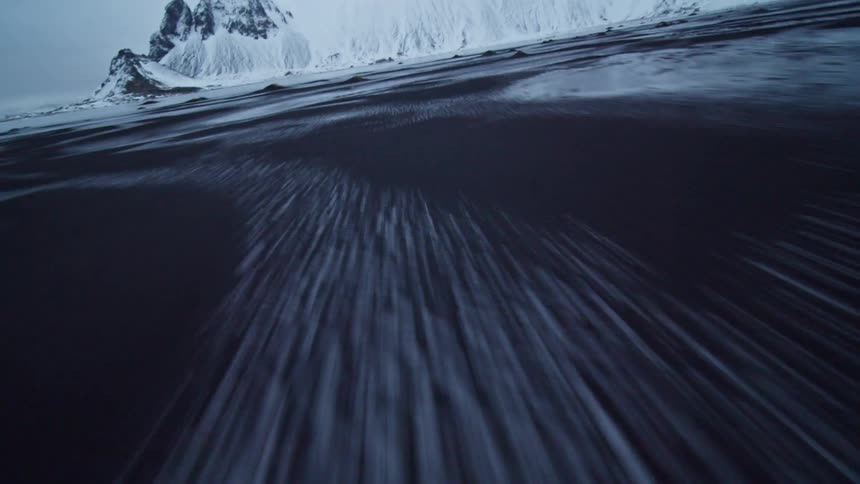 A dramatic low-angle aerial shot flies at high speed over a frozen black sand beach towards a majestic snow-covered mountain range in Iceland
