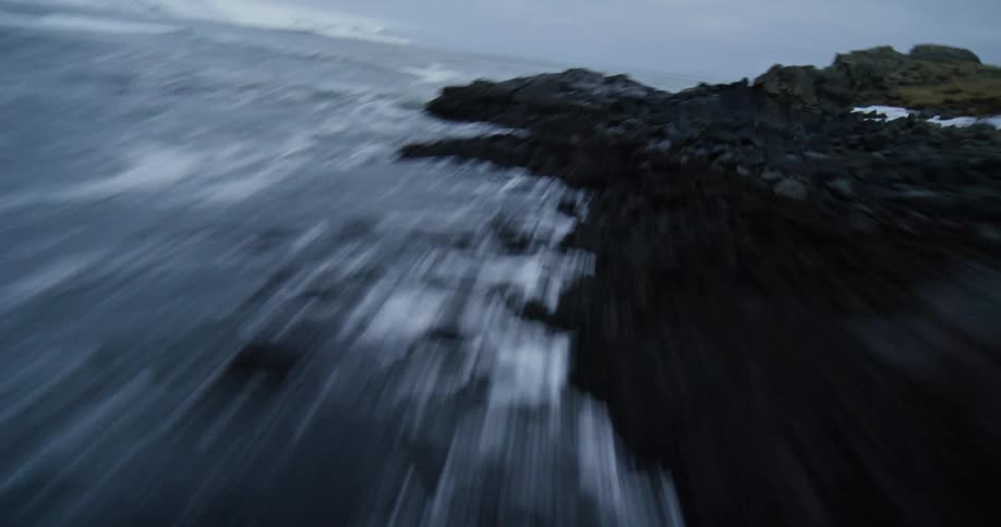 A thrilling FPV drone perspective flies at high speed along a dark sandy beach before soaring over the icy, rocky coastline on a bleak winter day