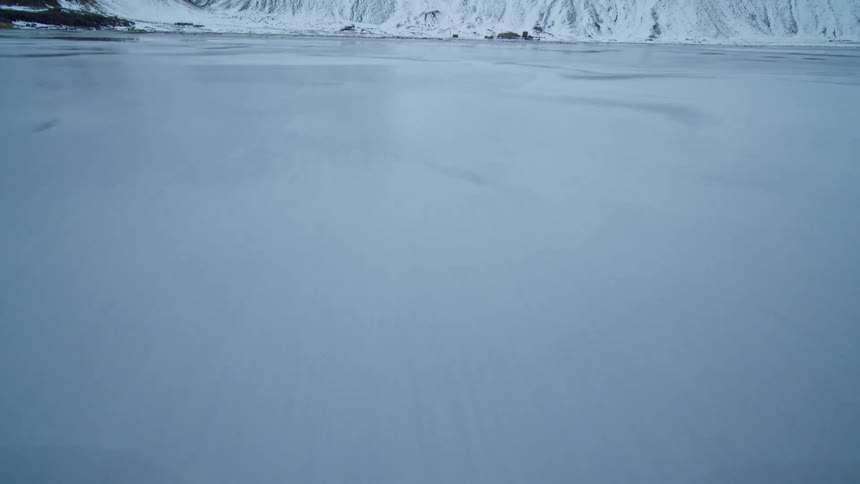 A fast-moving aerial shot skims low over a frozen, reflective black sand beach, with snow-covered mountains in the distance