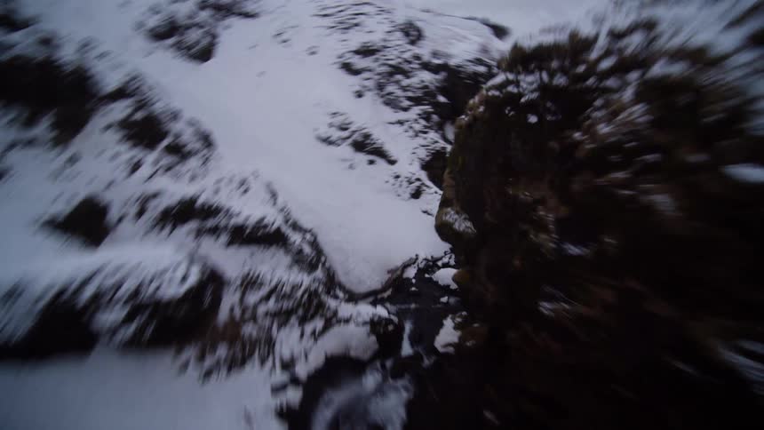 A thrilling first-person view drone flight dives down a steep, snow-covered canyon, following the winding river through the icy winter landscape