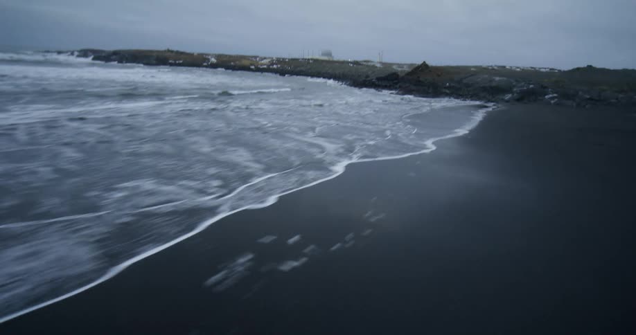 A dramatic, fast-paced aerial shot flies low over the white foam of waves on a black sand beach in a cold, snowy Icelandic landscape