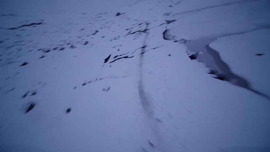 A first-person perspective glides quickly over a vast, snow-covered frozen lake, revealing a network of dark cracks in the ice