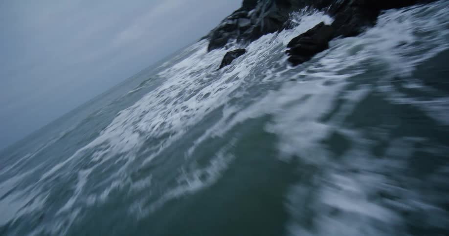 A dynamic FPV drone shot flies low and fast over a turbulent sea as waves crash against a rugged, snow-dusted rocky coastline on a bleak, overcast day