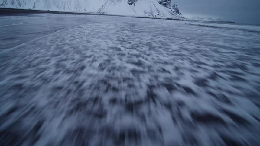 A dramatic low-angle shot captures the hypnotic motion of waves receding on a black sand beach in a frozen, winter landscape