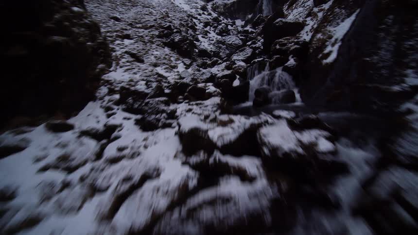 A fast-paced FPV drone shot soars up a winding river through a snowy canyon towards a dramatic waterfall
