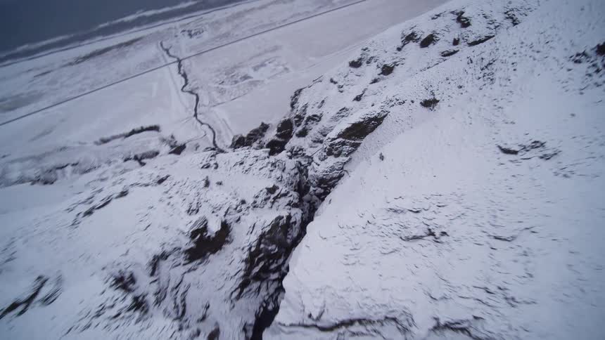 A thrilling FPV drone dive follows a winding river through a majestic, snow-covered canyon towards the distant ocean