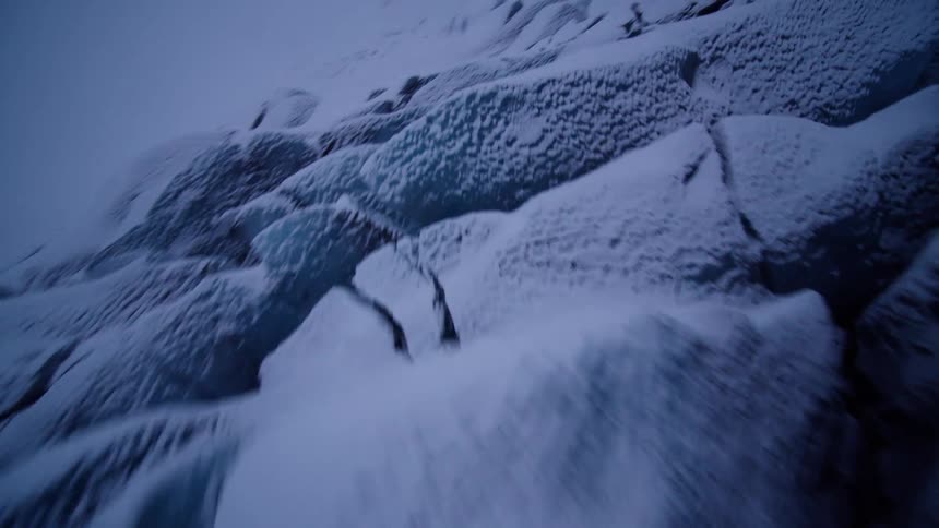 A dramatic FPV drone flight swoops low over the deep blue crevasses of a vast, snow-covered glacier on a foggy, overcast day