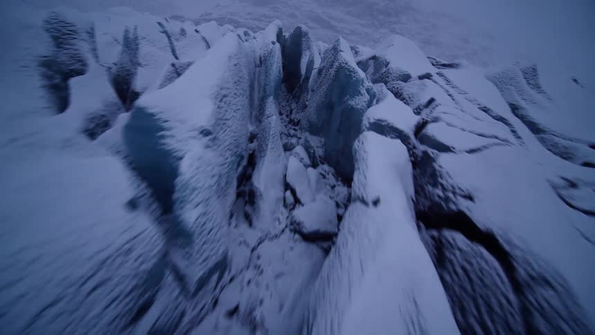 A fast-paced, first-person drone shot soars low over a snowy, frozen landscape and into the deep blue crevasses of a massive glacier