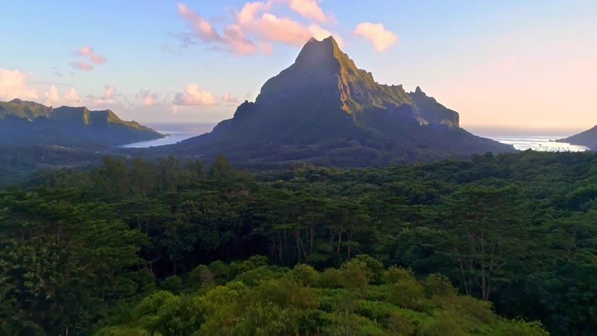 A drone flies past a woman on a hilltop towards an epic view of a majestic tropical mountain surrounded by a lush rainforest at sunset