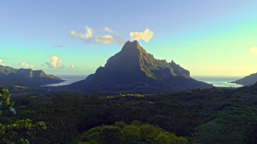 Aerial view slowly reveals a majestic mountain peak over a lush jungle on a tropical island at sunrise