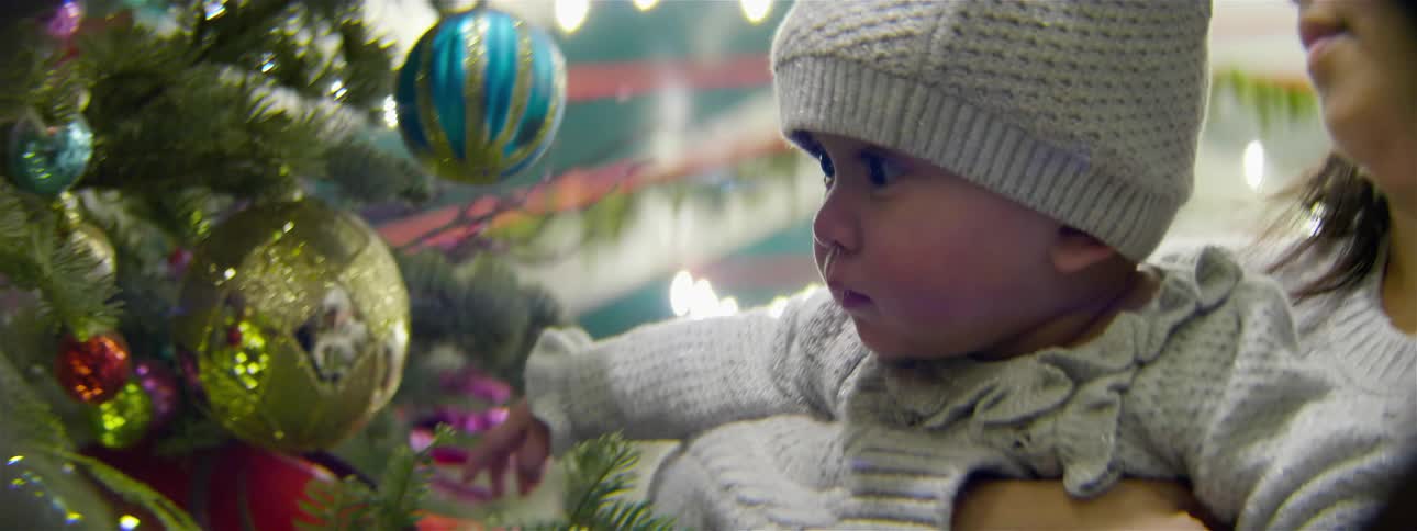 Baby held by woman looks at and touches ornament on christmas tree