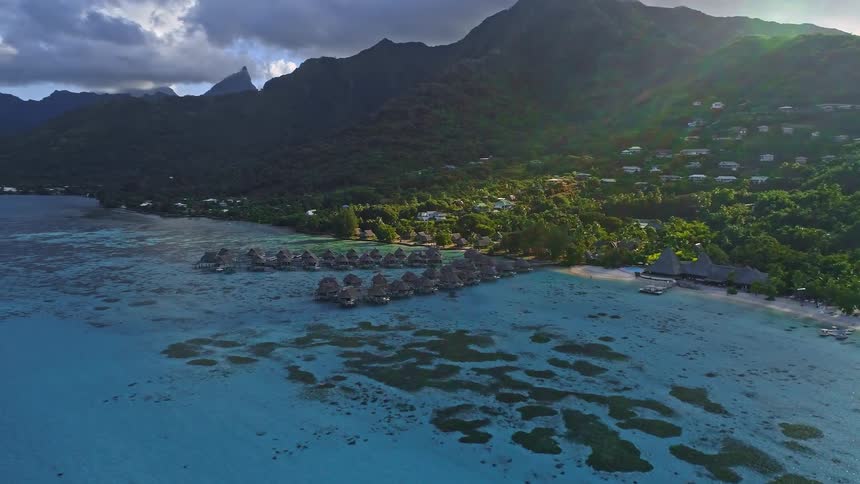 A breathtaking aerial view reveals a tropical island resort with overwater bungalows nestled in a turquoise lagoon at the foot of lush green mountains