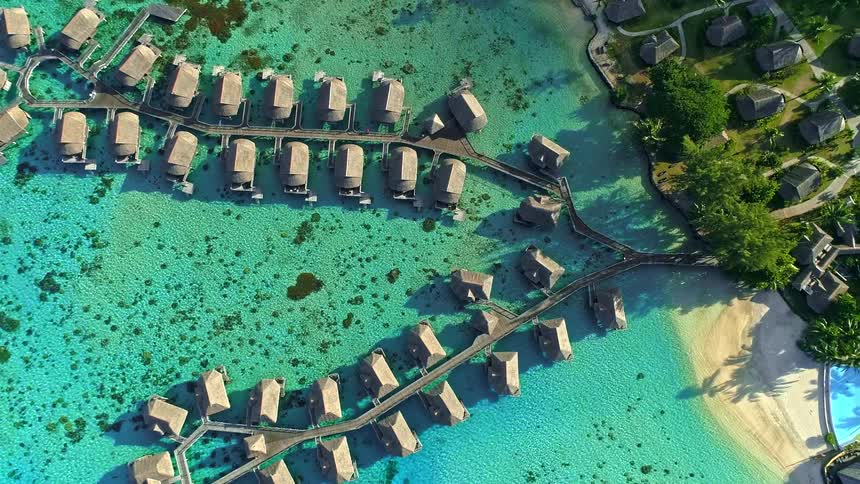 A stunning top-down aerial view slowly ascends over a luxurious tropical resort featuring overwater bungalows in a crystal-clear turquoise lagoon